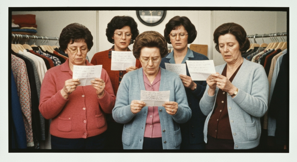 Six women in a clothing rack setting, reading small slips of paper, appearing serious and focused.